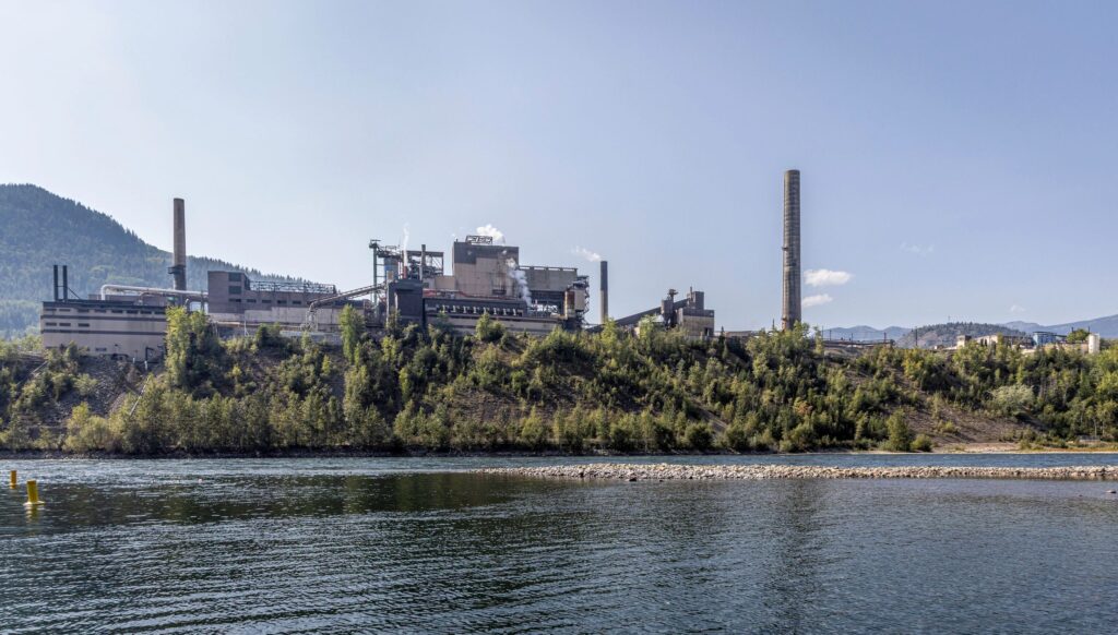 Industrial landscape across the Columbia river: a large smelter and factory complex with smokestacks on a hill, surrounded by trees, under a clear blue sky in Trail, British Columbia, Canada